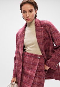 Red checkered blazer and skirt set in textured fabric, with a light beige turtleneck. Features accented buttons and a tailored fit.