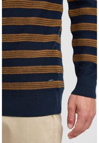 Pull à rayures bleu marine et moutarde avec un tissu texturé, des poignets côtelés et une petite étiquette de marque près de l'ourlet. Gros plan sur le bras et la main.