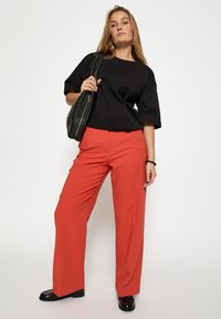 Schwarzes, kurzes T-Shirt mit lockerer Passform, kombiniert mit hochgeschnittenen, leuchtend roten weit geschnittenen Hosen. Das Modell trägt eine schwarze Schultertasche und glänzende Schuhe.