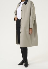 Lichtgrijze trenchcoat met zwarte kraag, voorzien van grote knopen en een losse fit. Gedragen over een wit topje en zwarte jeans. Gladde textuur.