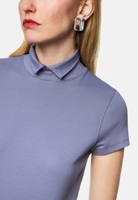 Helles lila Poloshirt mit kurzen Ärmeln und einem kleinen Kragen. Glatte Stofftextur und sichtbare Nähte. Metallische quadratische Ohrringe.