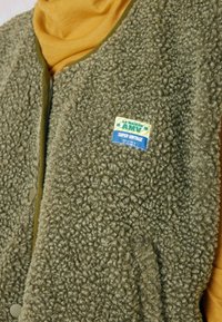 Fleece vest in olijfgroen met een textuurrijke, pluchen oppervlakte. Heeft een drukknoopsluiting en een kleurrijke tag met de tekst "LA MAISON AMV SUPER VINTAGE."