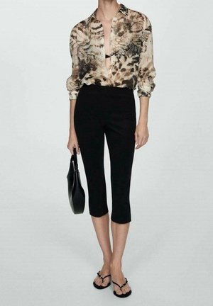 Frau trägt eine Bluse mit Leopardenmuster, schwarze kurze Hose, schwarze Sandalen und hält eine schwarze Handtasche vor einem einfarbigen Hintergrund.