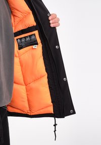 Zwarte pufferjack met een feloranje interieur, met een gestikt patroon en een ritzakken met onderhoudsinstructies.