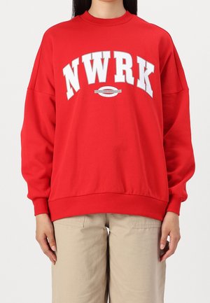 Personne portant un sweat rouge avec "NWRK" en grandes lettres blanches et un pantalon beige, montrée des épaules jusqu’aux hanches.
