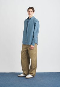 Svijetloplava košulja na dugme napravljena od pamuka, uparen je s širokim bež hlačama. Model nosi bijele tenisice, stoji pred neutralnom pozadinom.