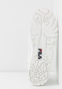 Semelle blanche de chaussure FILA montrant un motif texturé avec des motifs géométriques et manuels, ainsi que le logo FILA en noir et rouge au centre.