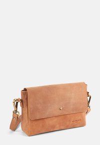 Tan leather crossbody bag with a fold-over flap, gold hardware, and a subtle embossed logo, featuring a removable strap and minimalistic design.