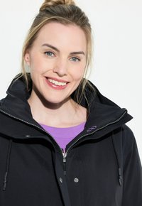 Svart vattentålig jacka med hög krage och framdragkedja, med en lila logotyp och dragsnören. Bärs över en lila skjorta.