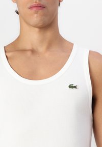 Hvit stripete tank topp med rund hals, med en liten grønn krokodillelogo på venstre side. Glatt stofftekstur.