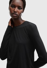 Top lungo nero a maniche lunghe con scollatura rotonda, realizzato in tessuto liscio, caratterizzato da una vestibilità rilassata e un design minimalista senza motivi visibili.