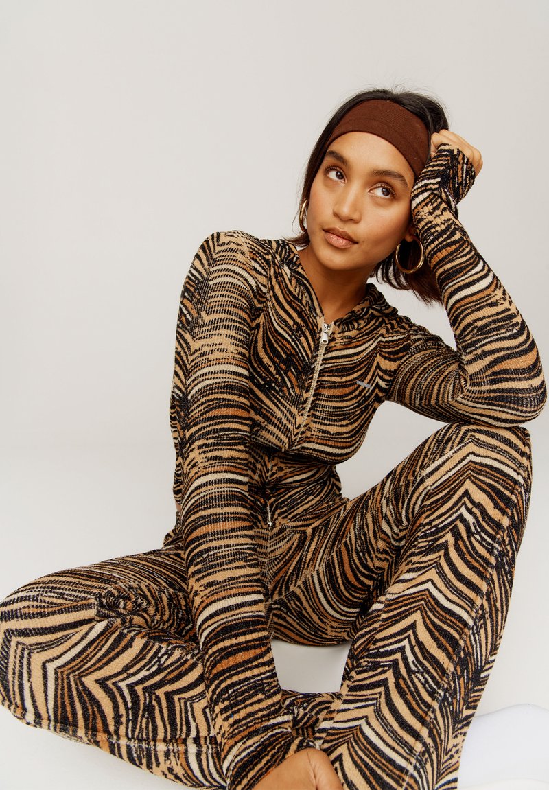 Beige, schwarze und braune, zebra-musterte, langärmlige Jumpsuit. Mit hohem Kragen und Reißverschluss. Glattes, strukturiertes Material.
