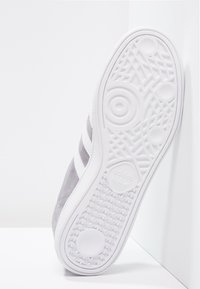 Szara podeszwa zamszowego buta sportowego z białą, fakturowaną gumową podeszwą zewnętrzną, wyposażona w trzy białe paski i okrągły wzór bieżnika.