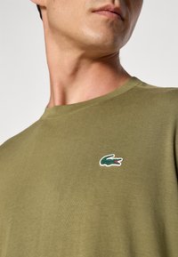 T-shirt de algodão verde-oliva com decote redondo e um pequeno logótipo de crocodilo bordado na área do peito esquerdo. Textura suave.