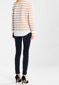 Haut à manches longues rayé en beige et blanc, superposé sur une chemise blanche, associé à un jean skinny noir et des escarpins noirs.