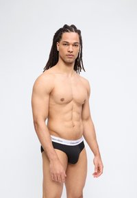 Μυώδης άνδρας με μακριά dreadlocks που φοράει μαύρα εσώρουχα Calvin Klein στέκεται μπροστά σε ένα απλό φωτεινό φόντο, κοιτώντας μπροστά.