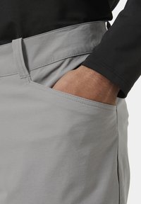 Šedé kalhoty se hladkým povrchem, s postranní kapsou a prošitým pasem, spárované s černou košilí s dlouhým rukávem.