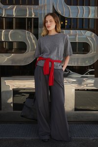 Maglietta grigia a maglia con maniche corte, tessuto rosso legato in vita e pantaloni larghi grigi scuri. Borsa nera posata di lato. Sfondo riflettente.