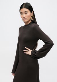 Ciemnobrązowa sukienka z długimi rękawami i wysokim dekoltem. Gładka tekstura z delikatnym rozciąganiem. Modelka nosi duże złote kolczyki w kształcie kół.
