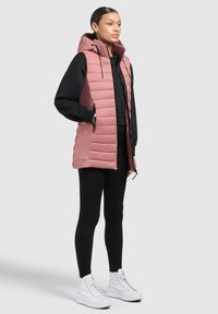 Rosa huva puffer väst med quiltat mönster, med dragkedjefickor, bärs över en svart tröja och svarta leggings, matchat med vita sneakers.