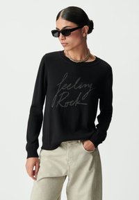 Maglione nero con grafica "feeling Rock" in filo argentato, abbinato a pantaloni larghi beige chiaro. Il modello indossa occhiali da sole e una collana d'oro.