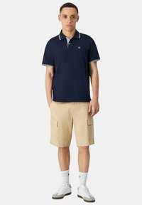 Polo shirt blu navy con colletto e polsini bianchi, abbinato a pantaloni cargo beige con tasche laterali, calze bianche e sneakers bianche.