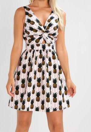 Hihaton vaaleanpunainen mekko, jossa musta ja keltainen ananaskuvio, solmittu edessä ja liehuva helma, päällä seisovalla henkilöllä.