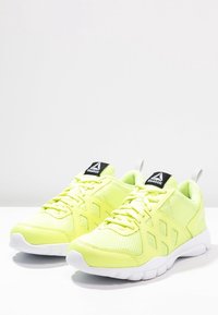 Helle, gelbe Sportschuhe mit einem Mesh-Obermaterial, Schnürverschluss, gepolstertem Sohlen und schwarzem Reebok-Logo auf der Lasche.