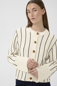 Tânără cu păr lung și castaniu, purtând un cardigan crem cu dungi verticale negre și nasturi mari, ajustând manșeta cu ambele mâini.