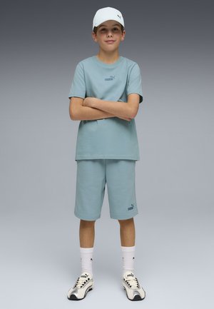 Niño de pie con los brazos cruzados, lleva una camiseta y pantalones cortos Puma azul claro, zapatillas Puma blancas, calcetines y una gorra blanca, fondo neutro.