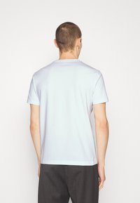 Man with short hair wearing a plain white short-sleeve t-shirt and dark gray pants, standing facing a white background.