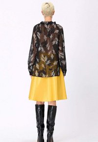 Blouse noire transparente avec un motif floral blanc, associée à une jupe en satin jaune et des bottes noires brillantes jusqu'aux genoux.