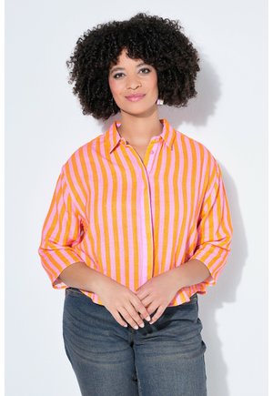 Kobieta o kręconych ciemnych włosach, ubrana w różowo-pomarańczową koszulę w paski zapinaną na guziki oraz ciemne dżinsy, stojąca na białym tle.