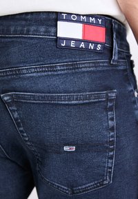 Tumši zili džinsi ir aprīkoti ar uzlīmi pie jostasvietas ar vārdiem "TOMMY JEANS" un taisnstūra formas sarkanu, baltu un zilu logo.