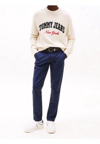 Jasnokremowy sweter z dzianiny z napisem „TOMMY JEANS New York” w granatowym i czerwonym kolorze; zestawiony z granatowymi chinosami i białymi trampkami z czarnymi akcentami.