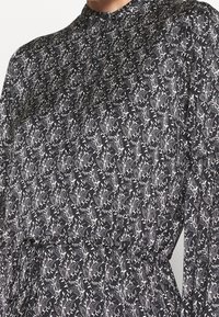 Robe en tissu noir avec motif floral blanc, col montant, manches longues et taille froncée, mettant en valeur un design texturé.