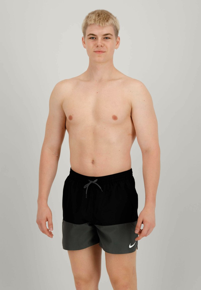 Costumi da bagno neri e grigi con cintura regolabile, logo Nike sul lato sinistro, tessuto leggero, indossati da un modello con capelli biondi corti.