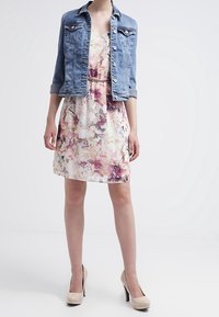 Jeansjacka med krage och framspringande knappar över en blommönstrad klänning. Klänningen har mjuka pastellfärger. Beiga högklackade skor.