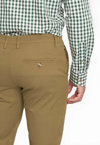 Pantalones color tostado con un bolsillo trasero y detalle de botón, combinados con una camisa de cuadros verde y blanca con puños abotonados.
