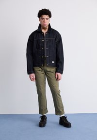 Giubbotto di jeans scuro con bottoni e cuciture dorate, abbinato a pantaloni verde oliva a gamba affusolata e scarpe nere con lacci. Design semplice e funzionale.