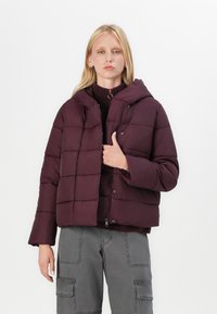 Giacca puffer bordeaux con cappuccio, dotata di chiusura a bottoni, design quadrato trapuntato e vestibilità comoda. Indossata sopra un maglione scuro.