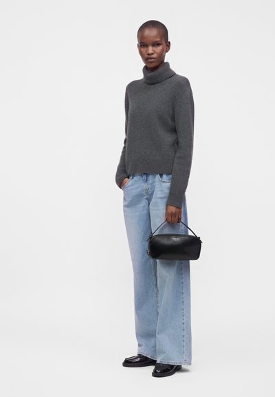 Femme portant un pull à col roulé gris foncé, un jean large bleu clair, des chaussures noires, tenant un sac à main noir d'une main.