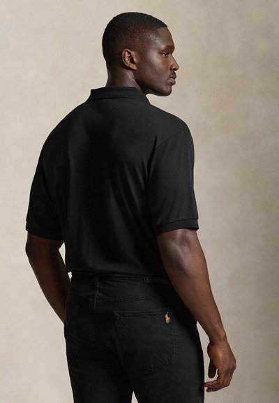 Mann trägt ein schwarzes Polohemd und schwarze Hose mit kleinem gelben Logo auf der Gesäßtasche, steht vor neutralem Hintergrund.