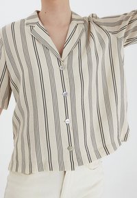 Camicia beige a maniche corte con strisce nere verticali, colletto a punta, due bottoni e una vestibilità rilassata. Tessuto morbido con leggera lucentezza.