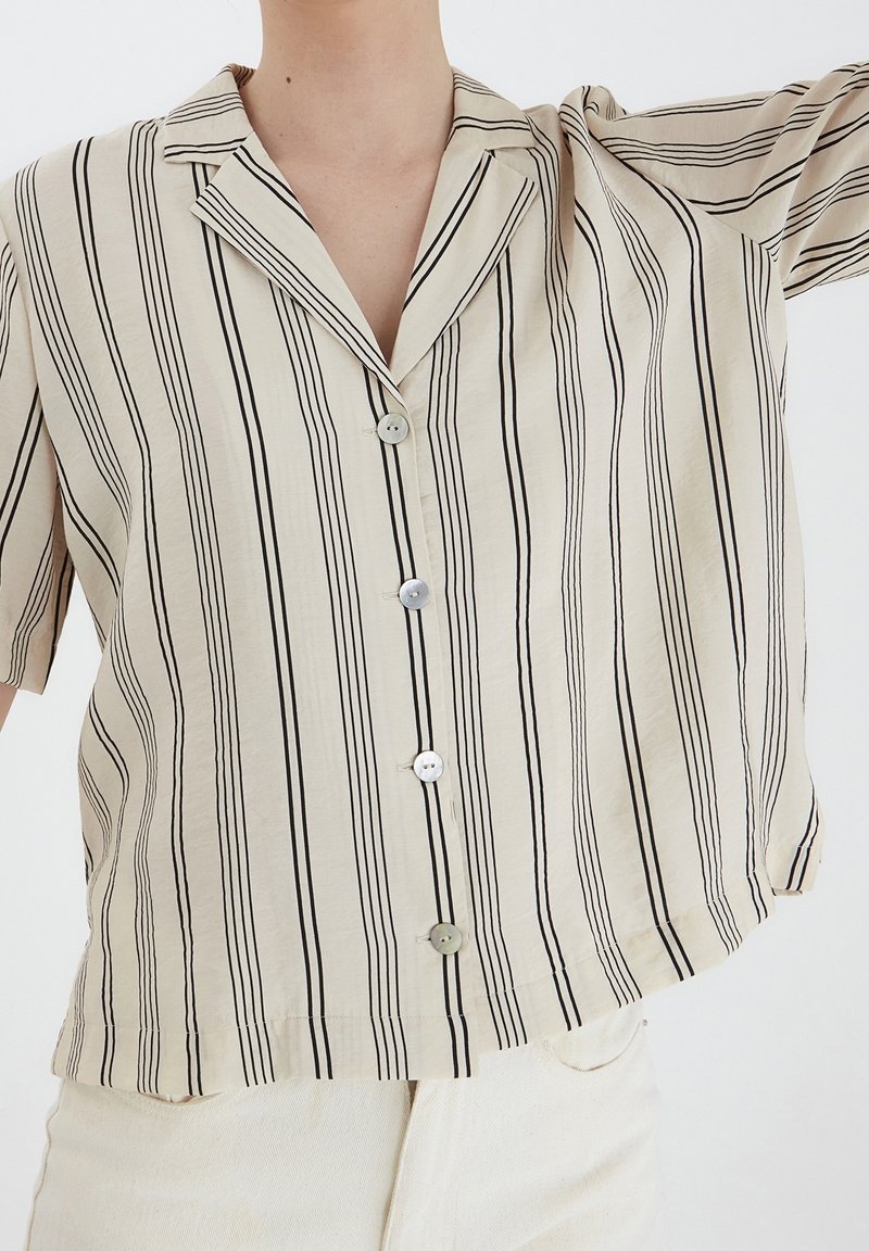 Camicia beige a maniche corte con strisce nere verticali, colletto a punta, due bottoni e una vestibilit�à rilassata. Tessuto morbido con leggera lucentezza.