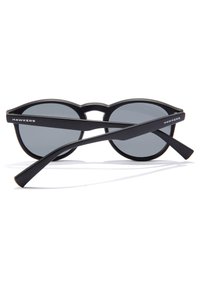 Occhiali da sole neri con lenti rotonde, design elegante e una caratteristica sagomatura nella parte superiore. Presentano aste sottili nere con testo del marchio in bianco.