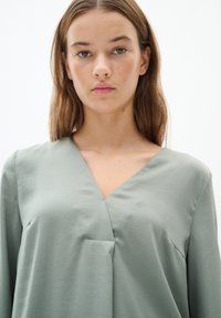 Blusa verde chiaro a maniche lunghe con scollatura a V e una sottile piega frontale, realizzata in un tessuto morbido e liscio. Design semplice senza motivi visibili.