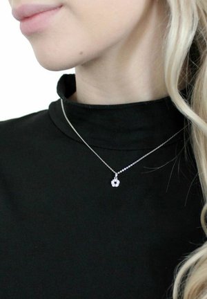 Silberne Halskette mit einem runden Anhänger, der mehrere kleine klare Steine aufweist, getragen über einem schwarzen Rollkragenpullover. Glatte, polierte Metalloberfläche.