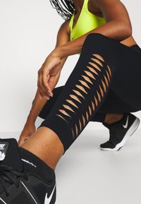 Osoba v černých leginách s šikmými výřezy, neonově žlutou sportovní podprsenkou a černými teniskami Nike, která se sklání na bílém podlaze.