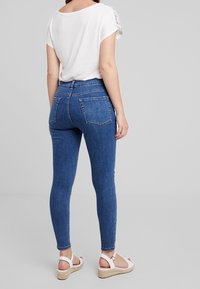 Donna vista di spalle che indossa jeans skinny blu, una maglietta bianca a maniche corte e sandali bianchi con cinturino, su uno sfondo neutro.